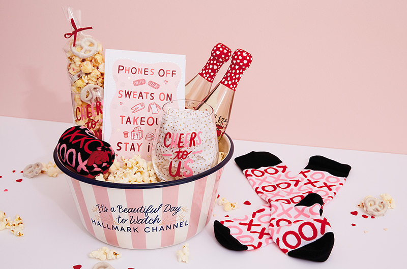 A Valentine's Day care package filled with gifts for a movie night in, including a Hallmark Channel popcorn bowl, wine glasses, cozy socks and a card.