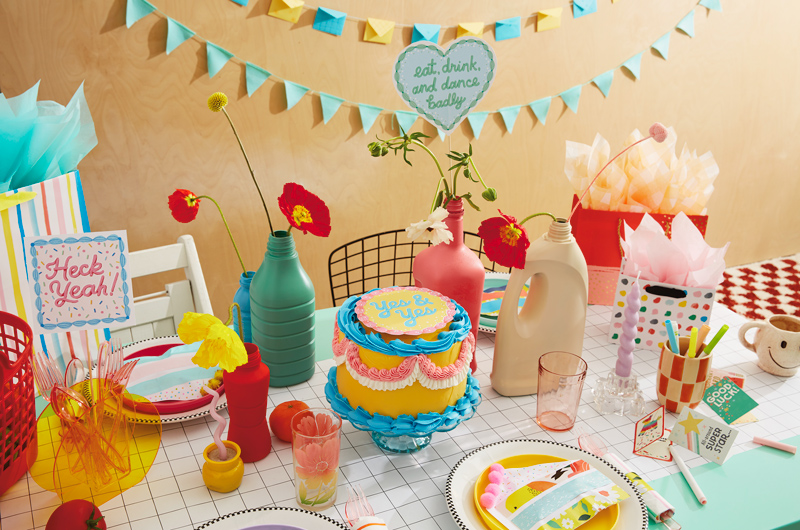 A table set with DIY party decorations, including a cake with a free printable cake topper, a mini card envelope garland, and cake favor pouches.
