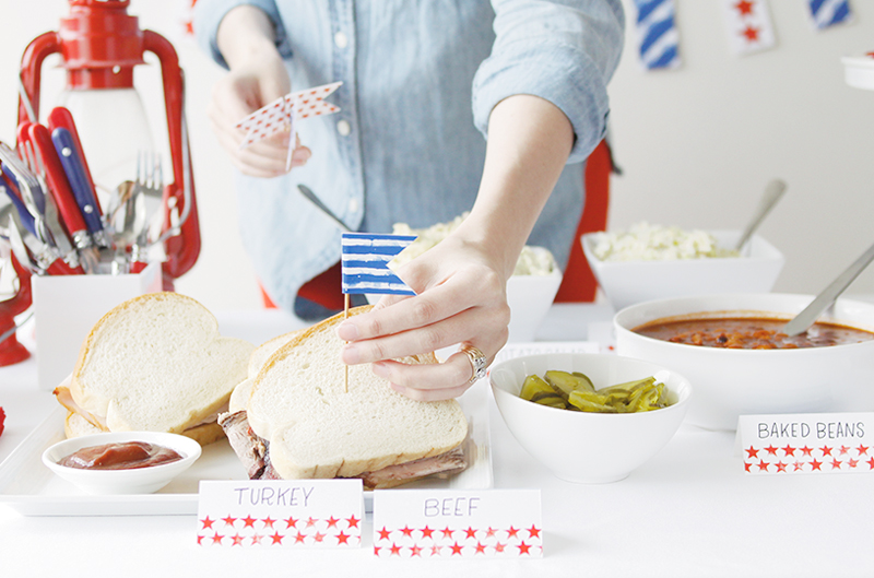 A hand placing a blue striped flag food marker into a sandwich, with tented food labels marked