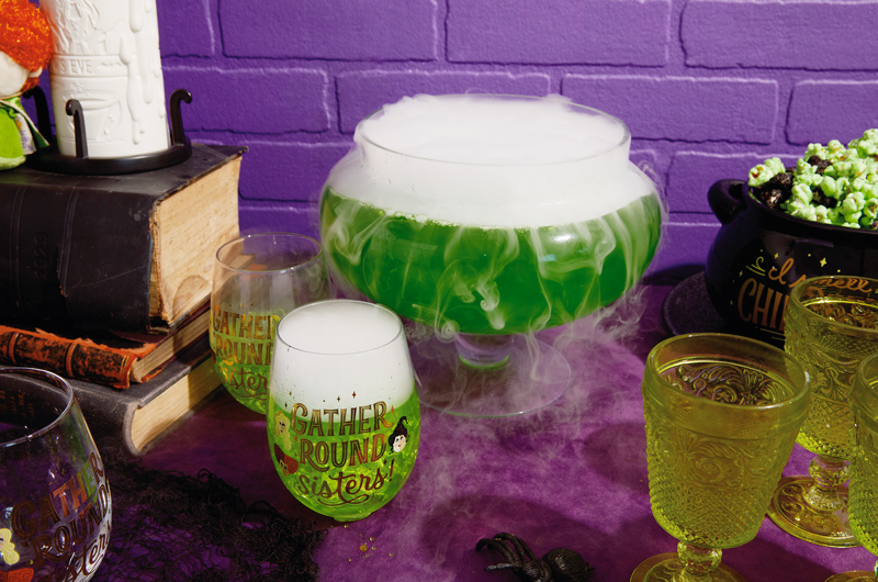 A punchbowl filled with a bright green drink; tendrils of mist spill over the sides. Nearby are Hocus Pocus stemless wineglasses that read,