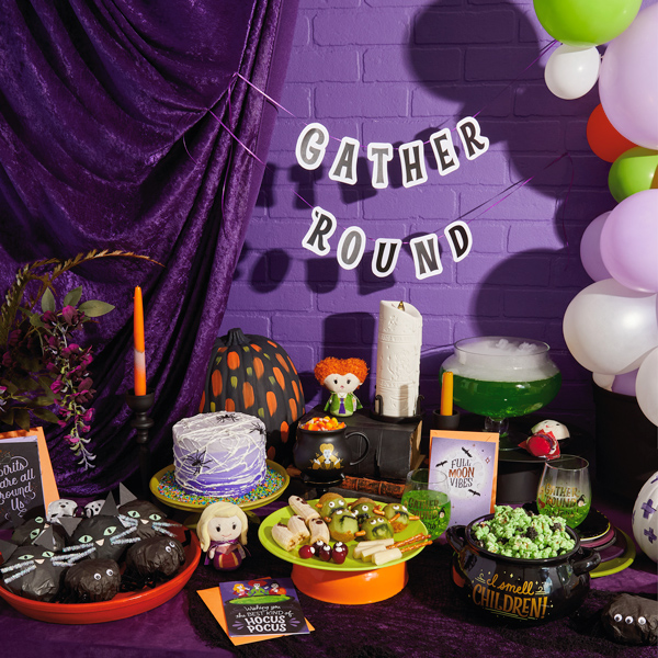 A Hocus Pocus party table, decorated with Hocus Pocus itty bittys, various Halloween treats, tissue paper popcorn balls shaped like black cat faces. Hung above it all is the free printable