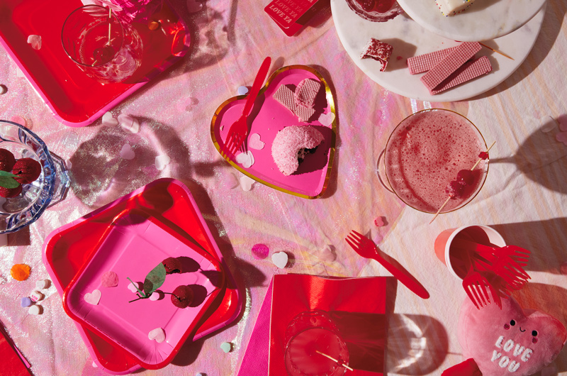 Looking down at a table set for a Valentine's Day party, with pink heart-shaped paper plates edged with gold foil and red and pink square paper plates. Cocktails and chocolate cherry cake truffles sit nearby.
