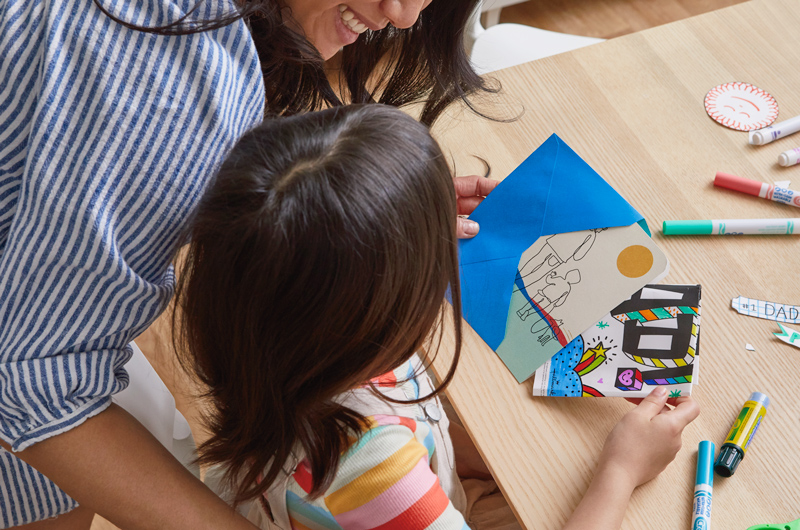 A woman smiles down at her young daughter as the little girl slides a completed free printable Father's Day coloring page into an envelope with his Father's Day card.