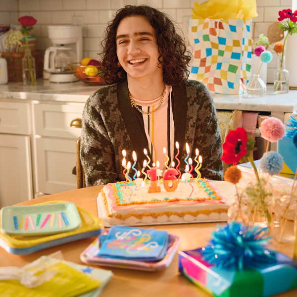 A teenage boy sits smiling at a kitchen table behind an iced birthday cake with a lit