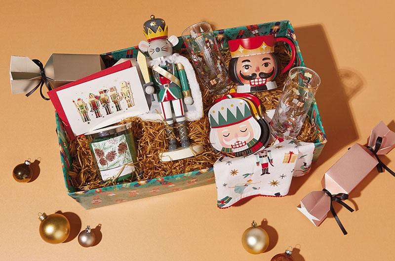 A Christmas care package filled with nutcracker-themed gifts, including nesting snack bowls, juice glasses, a mug, a mouse king nutcracker, a tea towel with a nutcracker pattern printed on it, and a nutcracker holiday greeting card.