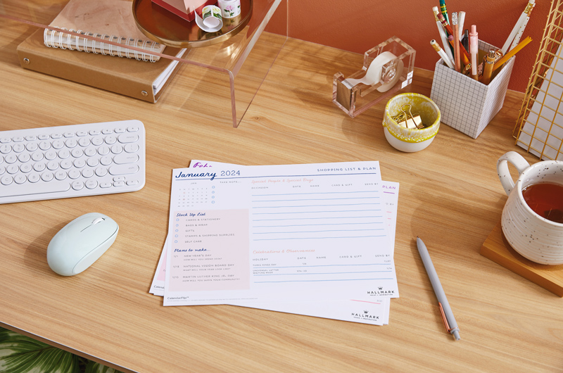 A printed January 2024 celebration planner page sitting on a home office desktop, surrounded by desk accessories.