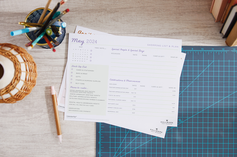 A printed May 2024 celebration planner page sitting on a home office desktop, surrounded by desk accessories.