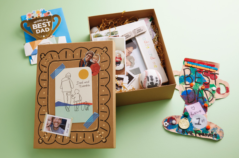A Father's Day memory box sits on a light green surface; the box is made of brown kraft cardboard; the top of the box is decorated with a Father's Day card taped down with blue and white striped washi tape, photos of a father and his toddler daughter smiling at the camera, and hand-drawn doodles surrounding everything in the style of a frame; inside the memory box are more photos, a baseball with a child's handprint on it in black ink, a white, ceramic frame with the words