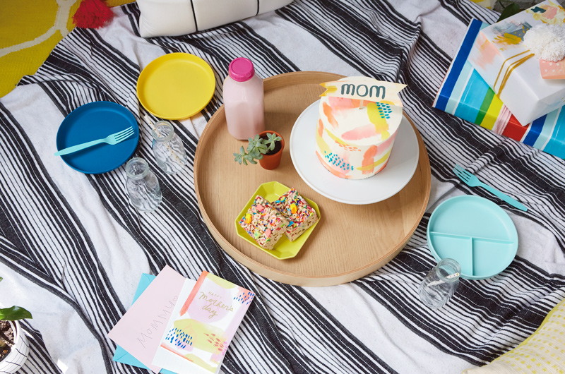 An indoor picnic setup for Mother's Day, with a black and white striped blanket on the floor; sitting on the blanket is a round wooden serving tray with a tall, round cake decorated with pale icing and a banner on top that reads,