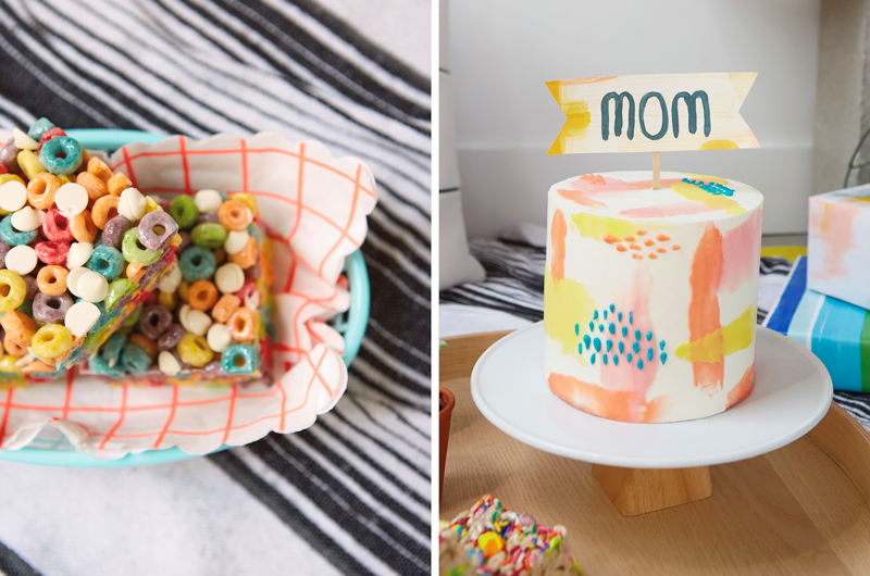 A black and white striped blanket with a basket of fruity breakfast cereal marshmallow squares sitting in it;a round wooden serving tray with a tall, round cake decorated with pastel icing in shades of orange, yellow, pink, blue and white, with a banner on top that reads,