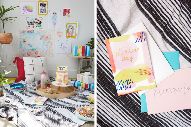 An indoor picnic setup for Mother's Day, with a black and white striped blanket on the floor; sitting on the blanket is a round wooden serving tray with a tall, round cake decorated with pale icing and a banner on top that reads,