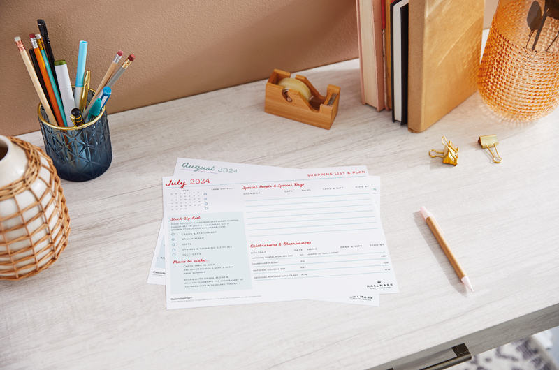 A printed July 2024 celebration planner page sitting on a home office desktop, surrounded by desk accessories.