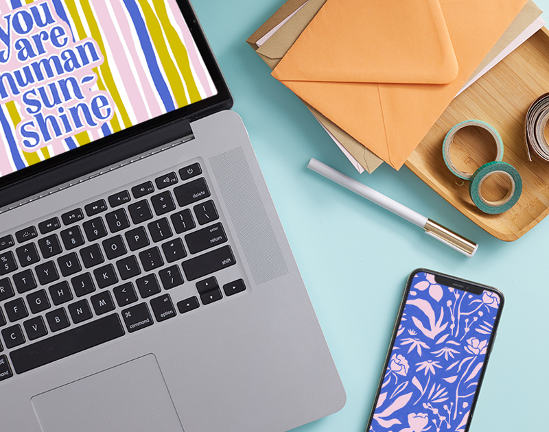 A laptop screen displaying the free August downloadable desktop wallpaper, with a mobile phone displaying the free downloadable mobile wallpaper lying nearby.