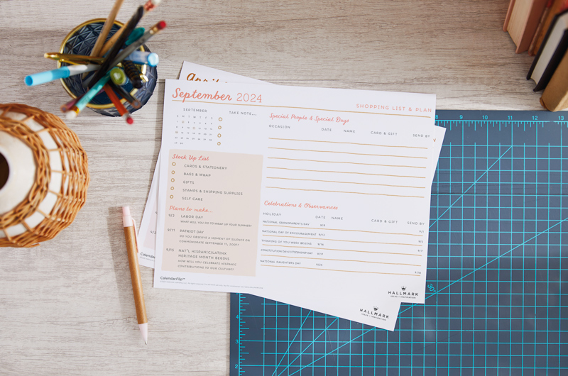 A printed September 2024 celebration planner page sitting on a home office desktop, surrounded by desk accessories.