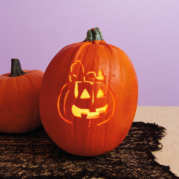 A pumpkin sits on top of a light wood table with a black mesh table runner, in front of a light purple walll; the pumpkin has been carved to feature a portrait of Peanuts Snoopy lying on top of a jack-o-lantern.