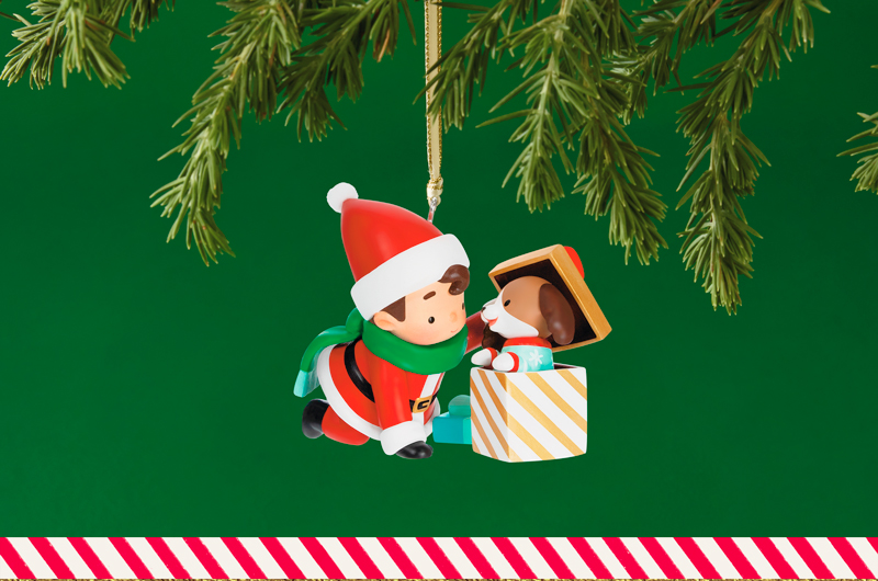 A Keepsake Ornament depicting Santa as a young boy; he is on his hands and knees, opening a gift box; popping out the top of the gift box is a puppy in a pair of blue pajamas covered in a pattern of white snowflakes.