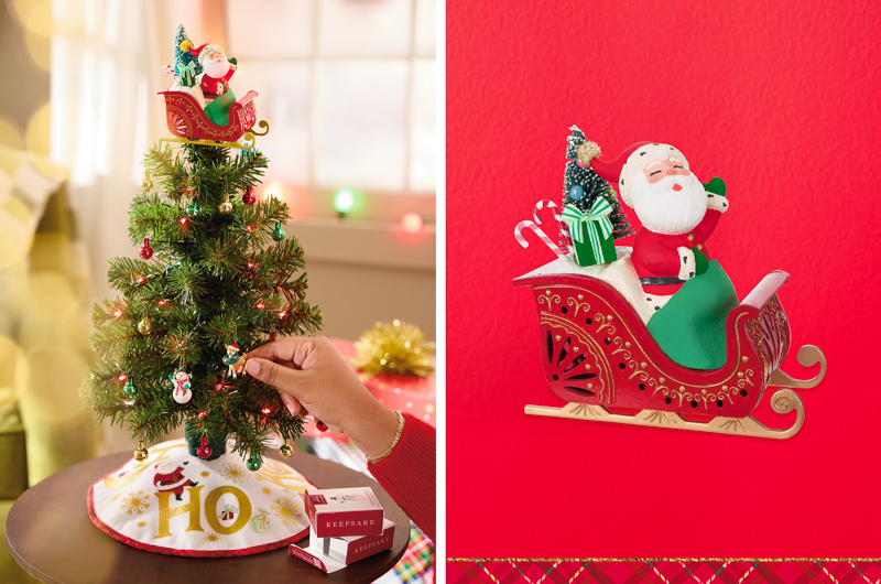 A mini tree covered in tiny red, green and gold ball ornaments and topped with a figurine showing a tiny Santa in a red and gold sleigh; the tree sits on a small, round wooden table and has a little tree skirt covering its base; the the skirt is white and reads