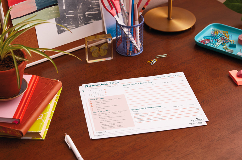 A printed version of the November celebration planner page sitting on a medium-toned wood desktop, surrounded by office supplies.